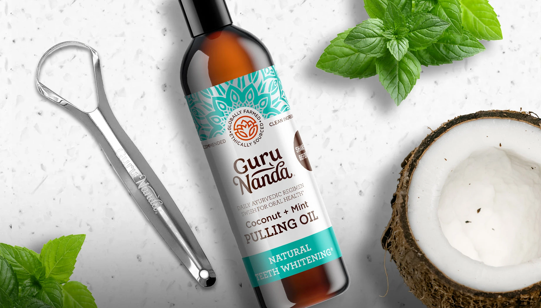 A bottle of GuruNanda pulling oil lying next to a steel tongue scraper, mint leaves and half a coconut